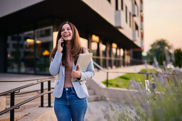 Fototapeta premium Confident Woman Talking on Phone and Holding Laptop Outside Modern Building