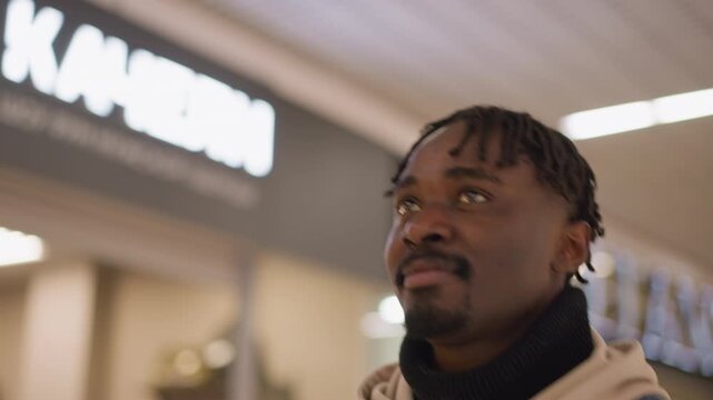 close up profile of young black man with dreadlocks wearing denim jacket and hoodie gazing upward in bright shopping mall corridor with blurred sign lights and reflective thoughtful expression