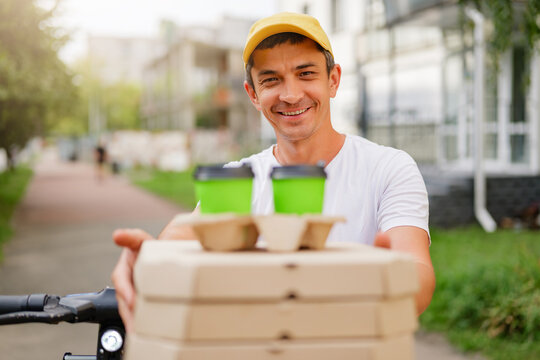 A cheerful delivery man on a scooter holds out a stack of pizza boxes and two green coffee cups. He's smiling directly at the camera on a sunny day, showcasing a friendly and reliable service.
