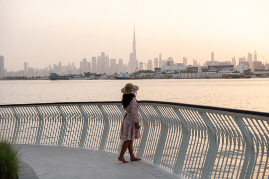 young woman wearing a hat and flowy dress exploring Dubai with Dubai skyline background, she is walking at the Creek harbor waterfront promenade at sunset
