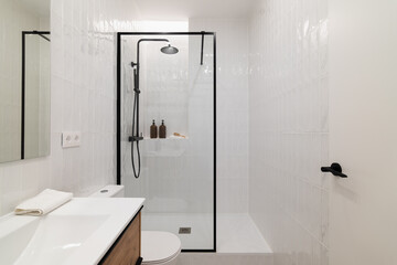 Modern bathroom with a glass shower enclosure, white tiles, and a wooden vanity. Minimalist design with toiletries on the shelf and a clean aesthetic.