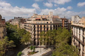 Urban landscape featuring historic buildings and green trees under a blue sky with clouds. A street is visible in the foreground.