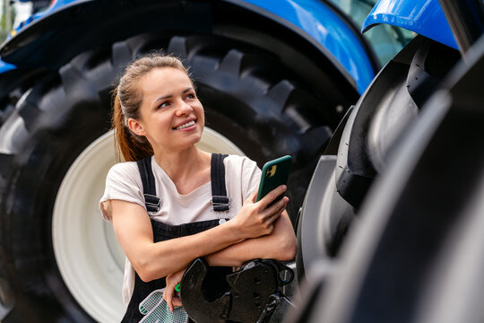 Female agricultural or construction machinery technician holding smartphone and looking at tractor - Powered by Adobe