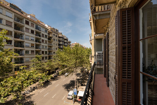 A sunny street view from a balcony in a city. Buildings line the street, with trees providing shade. Clear blue sky enhances the urban landscape.