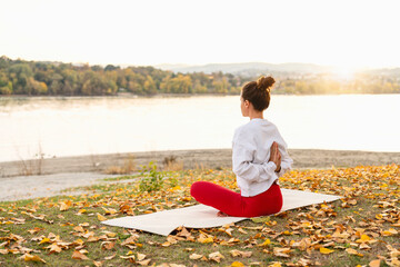 Rear view of woman meditating on yoga mat at riverside in autumn