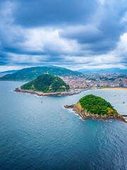 Obraz premium Scenic view of La Concha Bay in San Sebastian, Spain, featuring Santa Clara Island, Mount Urgull, dramatic clouds, lush hills, city skyline, and sandy beach.