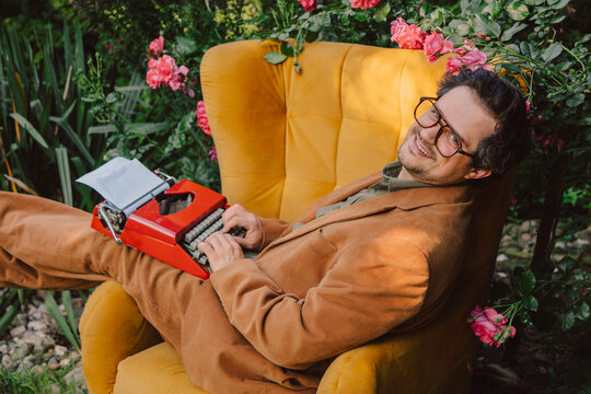 Stylish author relaxing in yellow chair with retro typewriter in blooming garden