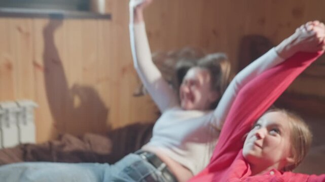 close up of joyful young women falling backward onto bed in warmly lit wooden room, smiling and enjoying carefree playful moment together while sunlight streams through window in cozy setting