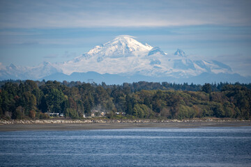 Snow-capped mountain looms over a serene waterfront landscape under a clear blue sky
