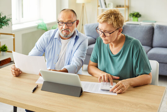  Senior couple reviewing bills and calculating expenses at home with tablet and paperwork. Financial planning moment. They focus on numbers and details. Calm, bright living room background.