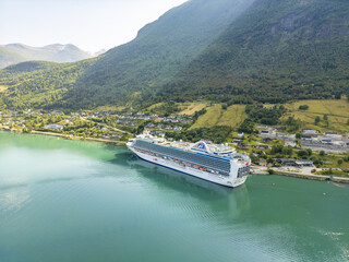 Olden, Norway - 21 July 2025: Aerial view of a colossal cruise ship nestled against the quaint village, reflecting in the fjord's turquoise waters beneath verdant mountains.