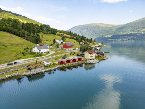 Aerial view of quaint houses nestled along the tranquil, deep blue fjord waters meeting lush green mountains under a bright, expansive sky, Olden, Vestland, Norway.