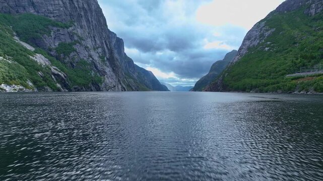 A drone glides over a fjord in Lysebotn, showcasing steep cliffs with greenery, calm reflective water, and dramatic peaks under a cloudy sky.