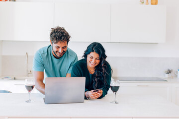 Couple using laptop and smartphone, drinking wine in kitchen