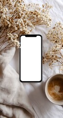 Flatlay of smartphone with blank screen, nestled amongst dried flowers, a beige blanket, and a cup of coffee on a white surface