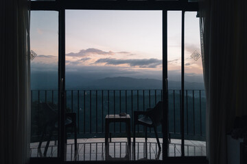 Two chairs and a table on a balcony overlooking a misty mountain range at sunrise, seen through a sliding glass door in Kandy, Sri Lanka