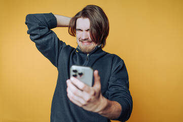portrait of young man expresses frustration while looking at his phone, with a visible wince and distressed expression on yellow background