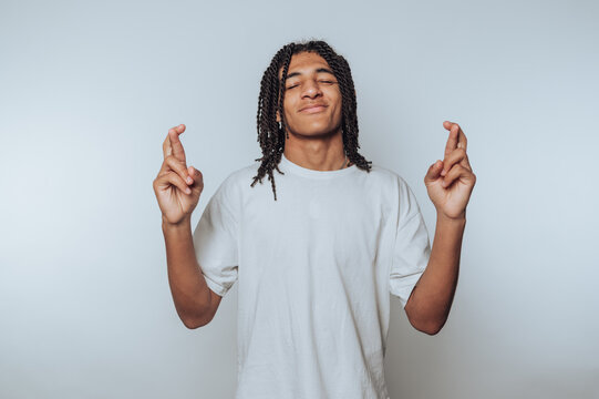 portrait of young man smiles while crossing his fingers, symbolizing hope and wishing for good luck on light background
