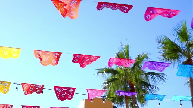 A low-angle view of colorful Mexican papel picado flags and palm trees against a clear blue sky, evoking a festive, cultural, and tropical atmosphere.