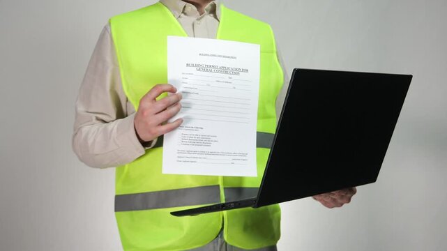 A construction specialist in workwear carefully examines important documents on a laptop. 