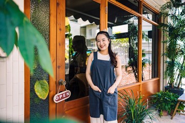 A young female coffee shop owner opens the door with a warm smile, welcoming customers, symbolizing...