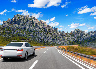 Naklejka premium A white sedan drives at high speed on a winding mountain road under a bright blue sky with scattered clouds.