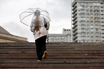 Rain in city, girl walking up with transparent umbrella the steps on city buildings background