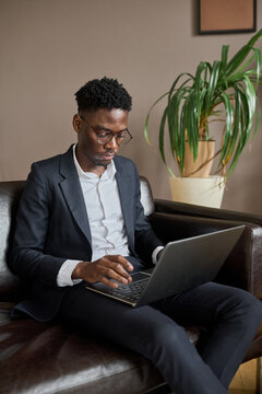 Vertical shot of young Black ceo wearing elegant suit sitting on sofa in modern office working on laptop
