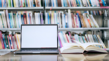 There is a laptop with a white screen and a book placed on the table in the library.
