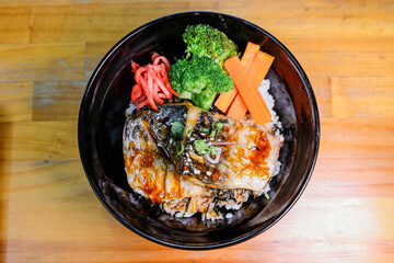 Close-up of Japanese grilled mackerel rice bowl with carrots, broccoli, pickled ginger, and soy glaze on wooden table.
