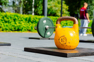 A yellow 16 kg kettlebell stands on a wooden stand on a sports ground. Green discs and a man in...
