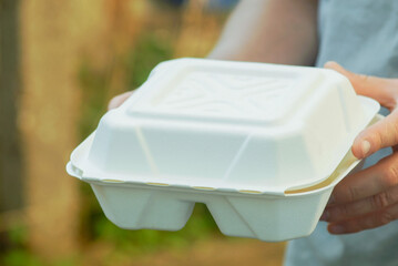 Close-up of male hand holding white takeaway food container outdoors. Сoncept of convenient lunch to go, ready meal packaging, street food delivery, quick service dining and portable meals