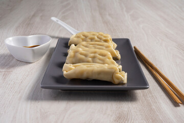 Four gyozas on a gray rectangular plate with wooden chopsticks, a spoon, and a bowl of soy sauce on a light wood surface.