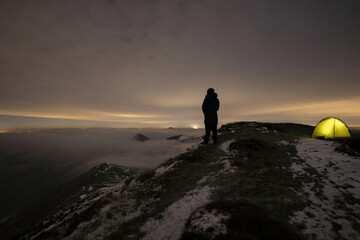Fototapeta premium A man standing on a hill at night beside his wild camping tent above a winter cloud inversion
