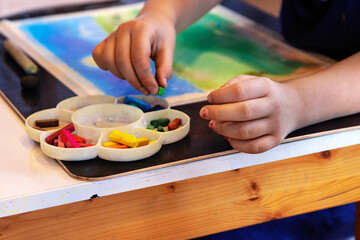 Closeup of a child’s hands selecting pastel chalks from a palette while working on a colorful drawing during the art lesson in school
