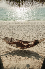 Woman in white bikini lying in a hammock between palm trees on a tropical beach, relaxing by the sea under the sunlight during summer vacation. 