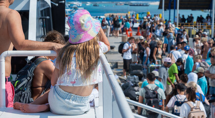a crowded pier with many people, boats, and luggage is viewed from the perspective of a child sitting on a ferry, wearing a sun hat.