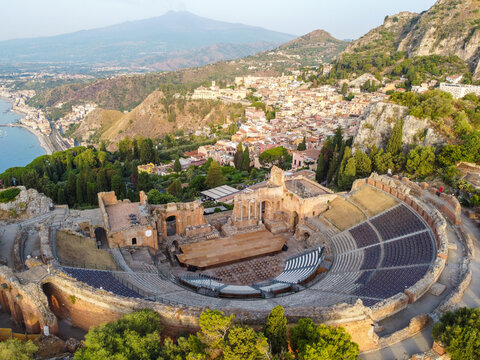 Aerial view of the ancient Teatro Greco's weathered stones meet the azure Ionian Sea, with Mount Etna's peak in the distance, Taormina, Sicilia, Italy.