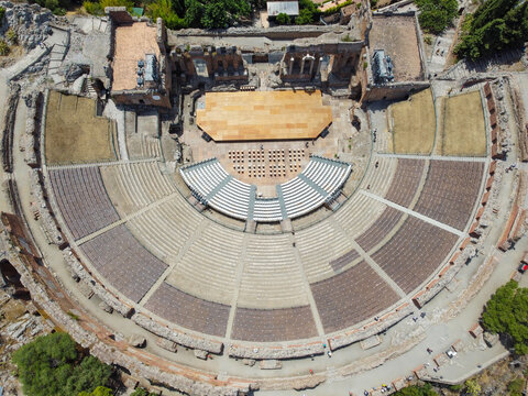 Aerial view of the ancient Teatro Greco, its stone tiers a warm contrast to the sun-drenched stage and surrounding greenery, Taormina, Sicilia, Italy.