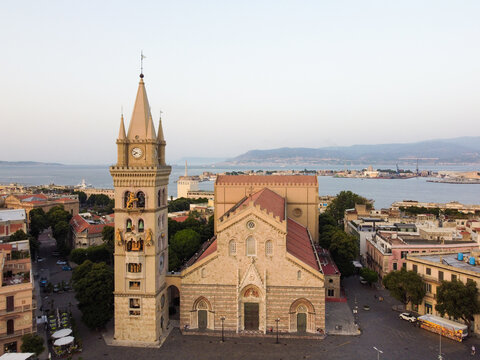 Aerial view of the Duomo's towering facade and intricate clock tower catch the golden light, framed by the azure sea and the city's rooftops, Messina, Sicily, Italy.