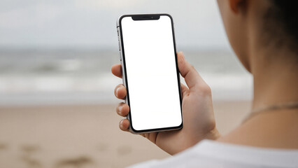 A person holds a smartphone with a blank white screen, with a blurry beach and ocean in the background. Ideal for showcasing mobile apps and digital content related to travel or outdoor themes.