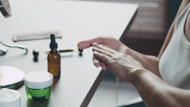 Handheld shot of mid-adult Caucasian woman applying peptide serum on face during anti-wrinkle beauty care routine in front of mirror at home