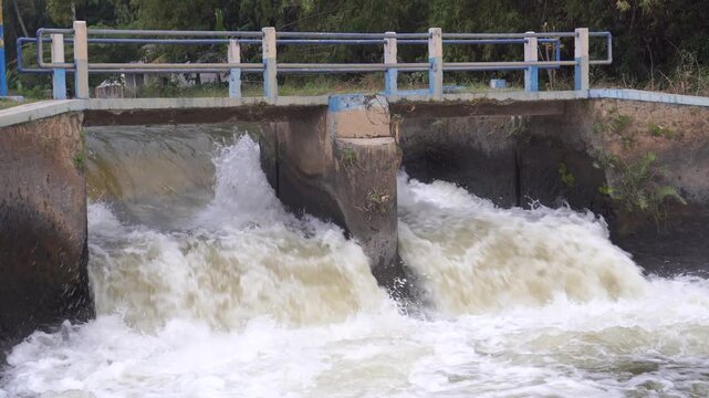 a powerful river being released through a concrete dam or spillway with two sluice gates. The video captures the immense force of the water as it cascades down, creating a turbulent, foamy river below