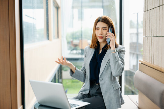 Confident Asian businesswoman using smartphone and laptop in a modern office, working remotely with smart tech. Stylish, professional