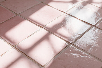 Warm light creates a geometric shadow pattern on an arrangement of pink-toned square tiles.