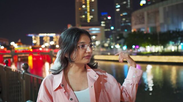 4K Portrait of confident Indian woman vlogging at Clarke Quay during night at Singapore city. Stylish female tourist filming a video and introducing tourist attractions. Holidays and travel concept.