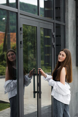 A girl with dark hair, wearing dark trousers and a white shirt, stands near a mirrored wall.
