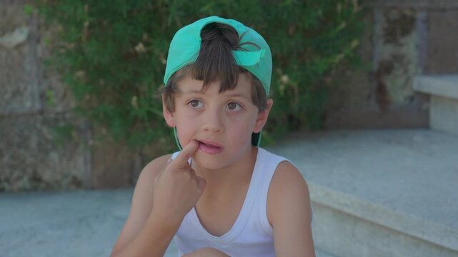 Young boy sitting outdoors on stone steps with hand near mouth wearing green baseball cap backwards and looking away with thoughtful expression