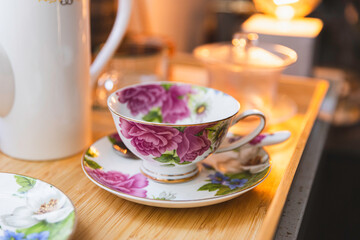 an elegant tea cup with floral decoration and white teapot on a wooden tray, warm yellow light in the background, afternoon tea set