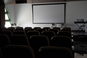 An empty lecture hall or auditorium with rows of seats facing a large blank projector screen. A perfect mock-up for presentations, corporate meetings, educational seminars, and movie screenings.